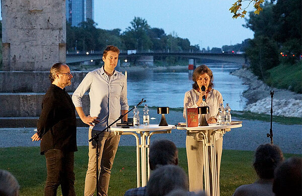 Lesung auf dem Schwal: Eine Frau spricht vor Publikum in ein Mikrophon, neben ihr am Stehpult lauschen zwei Männer, im Hintergrund die Donau