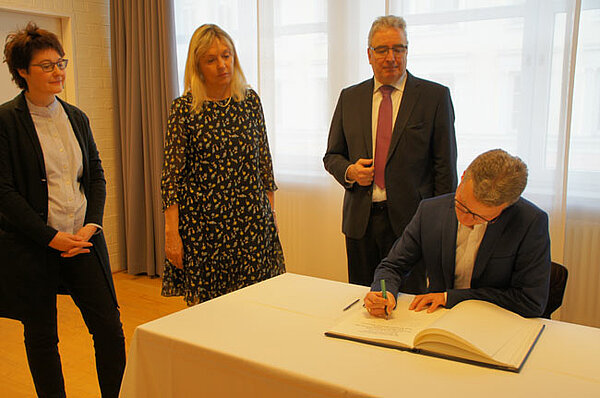 Staatsminister Bernd Sibler trägt sich in das Goldene Buch ein, Neu-Ulms Oberbürgermeister Gerold Noerenberg und zwei weitere Personen stehen daneben und sehen ihm dabei zu.