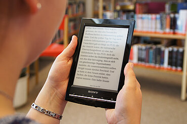 Blick über die Schulter einer Frau, die in einem e-Book-Reader liest, im Hintergrund Bücherregale