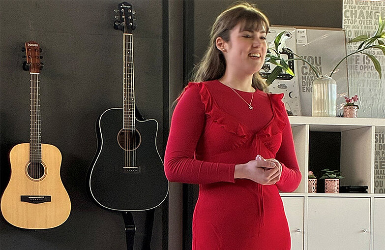 Eine junge Frau singt, hinter ihr hängen zwei Gitarren an einer Wand.