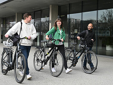 Drei junge Personen laufen nebeneinander und schieben je ein Fahrrad.