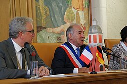 Neu-Ulms Oberbürgermeister und Bois-Colombes Bürgermeister sitzen während der offiziellen Zeremonie an einem Tisch, Bois-Colombes Bürgermeister spricht ins Mikrophon