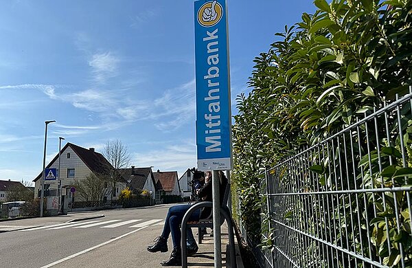 Sitzbank an einer Straße, daneben ein Hinweisschild mit der Beschriftung "Mitfahrbank"
