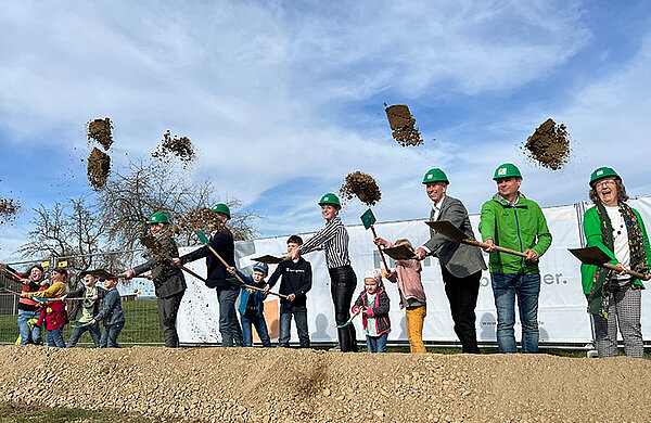 Erwachsene und Kinder werfen beim Spatenstich mit einem Spaten Erde in die Luft