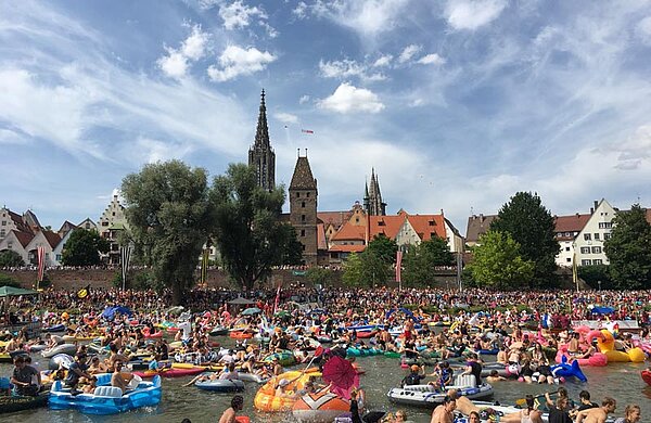 Foto vom „Nabada“: Tausende Menschen baden auf Flößen, Luftmatratzen und kleinen Booten die Donau hinunter. Im Hintergrund die Ulmer Skyline mit dem Ulmer Münster.