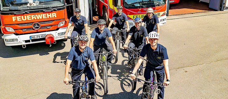 Kolleginnen und Kollegen der Feuerwehr stehen mit Fahrrädern vor Feuerwehr-Fahrzeugen