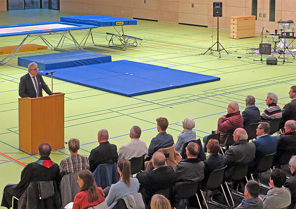 Man sieht Oberbürgermeister Noerenberg am Podium, das in der Turnhalle aufgestellt ist, wie er seine Eröffnungsrede vor Publikum hält.