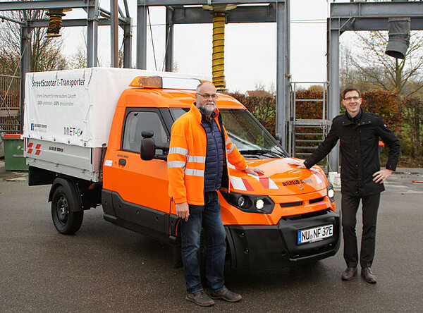 Das orangene Elektroauto mit Planenaufbau im Mittelpunkt des Bildes, vor dem Fahrzeug stehend zwei Personen; im Hintergrund Silos des Baubetriebshofes