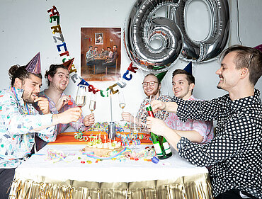 Fünf Personen mit Partyhüten sitzen an einem Tisch und stoßen mit Sektgläsern an, hinter ihnen an der Wand hängen zwei Luftballons in Form der Zahl 60.