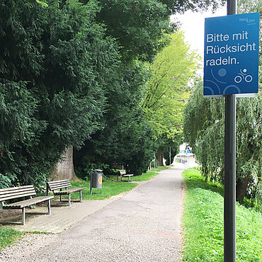 Blaues Schild mit der Aufschrift "Bitte mit Rücksicht radeln" am Neu-Ulmer Donauuferweg
