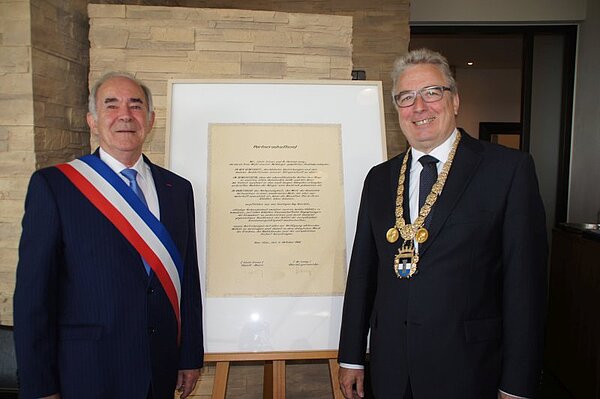 Bois-Colombes Bürgermeister Yves Revillion und Neu-Ulms OB Gerold Noerenberg stehen vor dem eingerahmten Partnerschaftseid von 1966 und lachen in die Kamera.