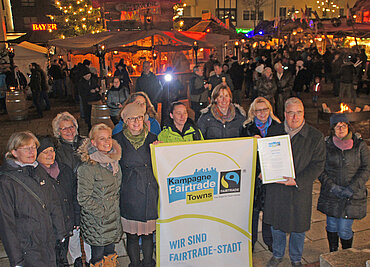 Zehn Personen stehend mit Blick in die Kamera, in der Mitte von ihnen ein Banner mit dem Logo "Kampagne Fairtrade Towns",  im Hintergrund Besucher und Marktstände des Mittelalterlichen Weihnachtsmarktes auf dem Neu-Ulmer Rathausplatz