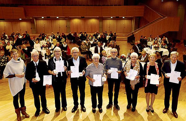 Gruppenfoto mit den Geehrten sowie der Oberbürgermeisterin, im Hintergrund sitzen weitere Gäste der Festveranstaltung an Tischen zusammen.