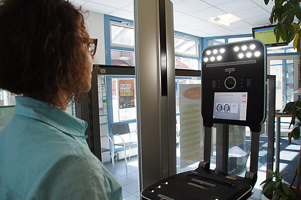 Foto von einer jungen Frau bei der Anwendung des Speed Capture-Terminals: Blick über die Schulter der Frau, die in die Kamera des Gerätes blickt; am Bildschirm werden zwei Passbilder von ihr angezeigt.