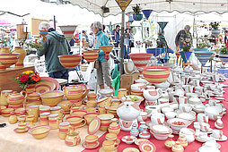 Marktstand mit bemalten und lasierten Keramik- und Töpferwaren wie Geschirr und Vasen, im Hintergrund Marktbesucher und Händler