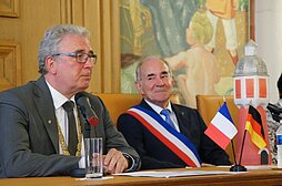 Neu-Ulms Oberbürgermeister und Bois-Colombes Bürgermeister sitzen während der offiziellen Zeremonie an einem Tisch, auf dem ein Mikrophon und eine Ständer mit Deutschland und Frankreich-Fahne steht