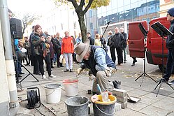 Der Künstler Gunter Demnig während der Stolpersteinverlegung am Boden kniend beim Arbeiten mit einer Spachtel, im Hintergrund Besucher der Gedenkveranstaltung