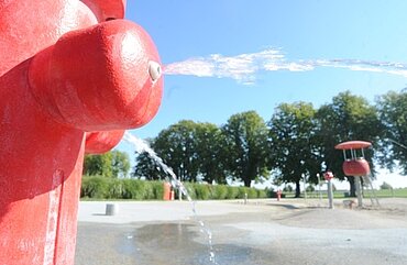 links groß im Bild ein großer roter Wasserhydrant, aus dem Wasser herausspitzt, im Hintergrund der Wasserspielplatz und Bäume