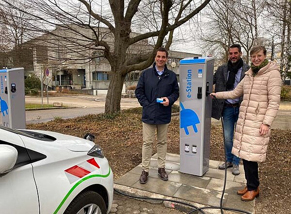 Oberbürgermeisterin Katrin Albsteiger steht an einer Strom-Ladesäule und lädt mit einem Ladekabel ein Elektroauto auf. Neben ihr die beiden Geschäftsführer der Stadtwerke und des Edwin-Scharff-Hauses.
