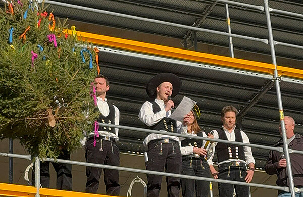 Mehrere Zimmermänner stehen auf einem Baugerüst, neben ihnen der Richtbaum