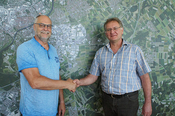 Der alte und der neue Leiter des Neu-Ulmer Baubetriebshofes stehen vor einer geografischen Landkarte von Neu-Ulm und reichen sich die Hand.