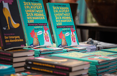 Ein Tisch mit Bücherstapel, unter anderem mit dem Buch "Der Hahn erläutert unentwegt der Henne, wie man Eier legt" von Ella Carina Werner.
