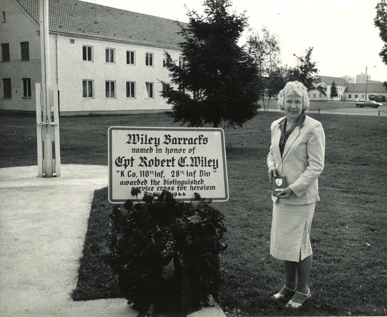 Robert Wileys Schwester neben einer Tafel mit der Aufschrift "Wiley Barracks - named in honor of Captain Robert C. Wiley"