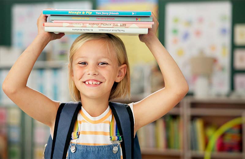 Ein Mädchen steht in einem Klassenzimmer und hält einen Stapel Bücher über den Kopf.