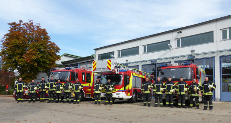 Gruppenfoto: Die Mitarbeitern der Feuerwehr-Wache in Feuerwehr-Montur vor ihren Einsatzfahrzeugen