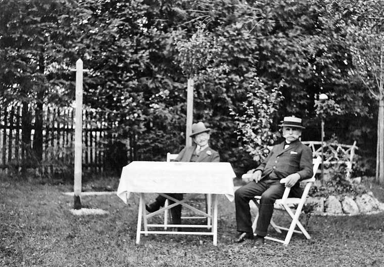 Zwei Männer mit Hut sitzen auf Klappstühlen an einem Tisch im Garten
