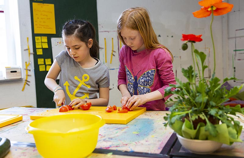 Zwei Kinder stehen an einem Tisch und schneiden auf einem Brettchen Tomaten in kleine Stücke.