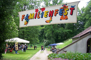 Ein Banner mit der bunten Aufschrift "Familienfest" im Eingangsbereich zu einem Park, im Hintergrund Spielstationen und Besucher 