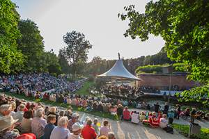Besucher eines Open-Air-Konzertes im Neu-Ulmer Stadtpark sitzen rund um die überdachte Veranstaltungsbühne