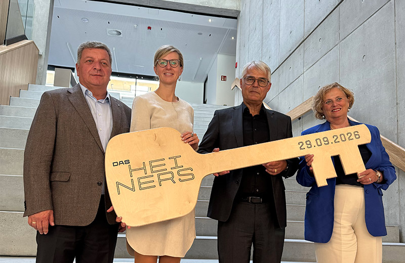 Vertreter von Stadt, Ministerium und beteiligten Partnern halten bei der Übergabe einen symbolischen großen Schlüssel mit der Aufschrift "Das Heiners".