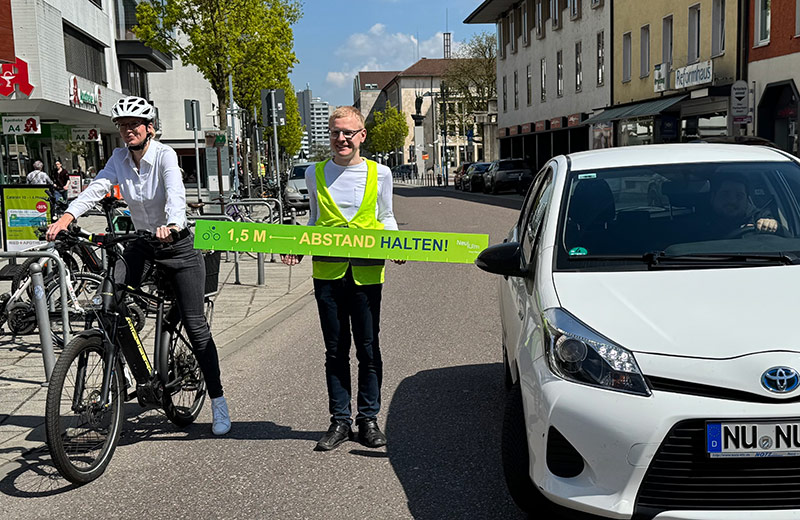 Oberbürgermeisterin Katrin Albsteiger auf dem Fahrrad am Fahrbahnrand. Sie wird von einem Auto überholt. Der Radverkehrsbeauftragte steht mit einem langen Maßband mit der Aufschrift "1,5 Meter Abstand halten" zwischen Fahrrad und Auto.