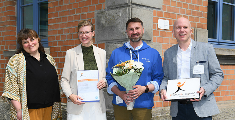 Vier Personen, eine mit einem Blumenstrauß und zwei andere mit Urkunde und Plakette Auszeichnung, blicken in die Kamera.