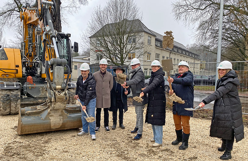 Vertreter von Stadt und Baufirma stehen beim Spatenstich neben einem Bagger und werfen mit einem Spaten Erde in die Luft.