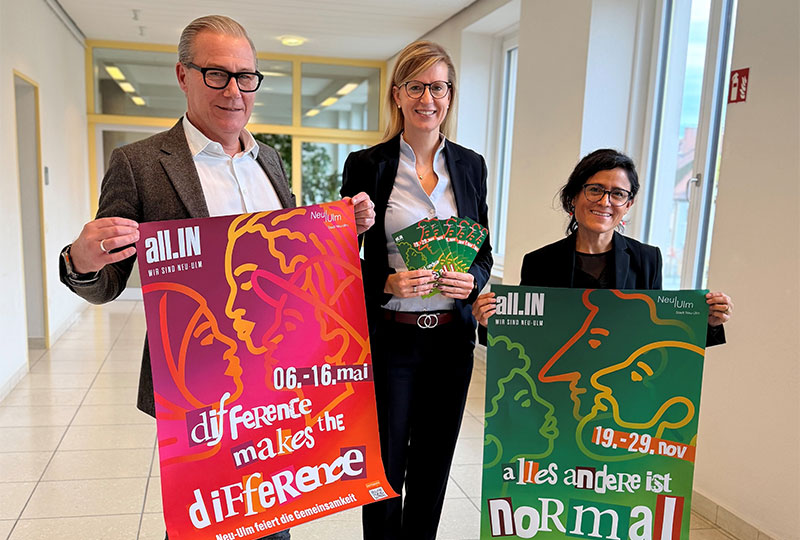 Die drei Personen halten Plakate und Flyer zur Veranstaltungsreihe all.IN vor die Kamera.