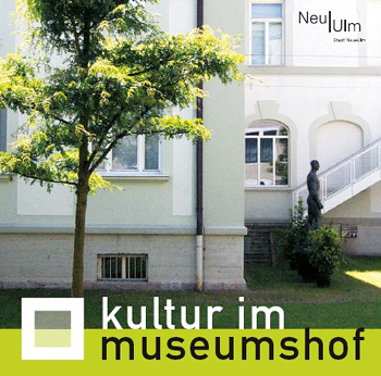 Bild vom Innenhof des Edwin Scharff Museums mit einem Baum, im Hintergrund die Museumsaußenfassade, unter dem Bild der Schriftzug Kultur im Museumshof